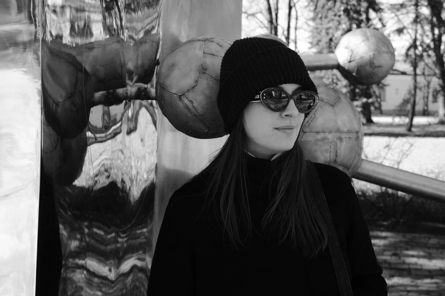 Black and white photo of a young woman in black hat and sunglasses next to a highly reflective object