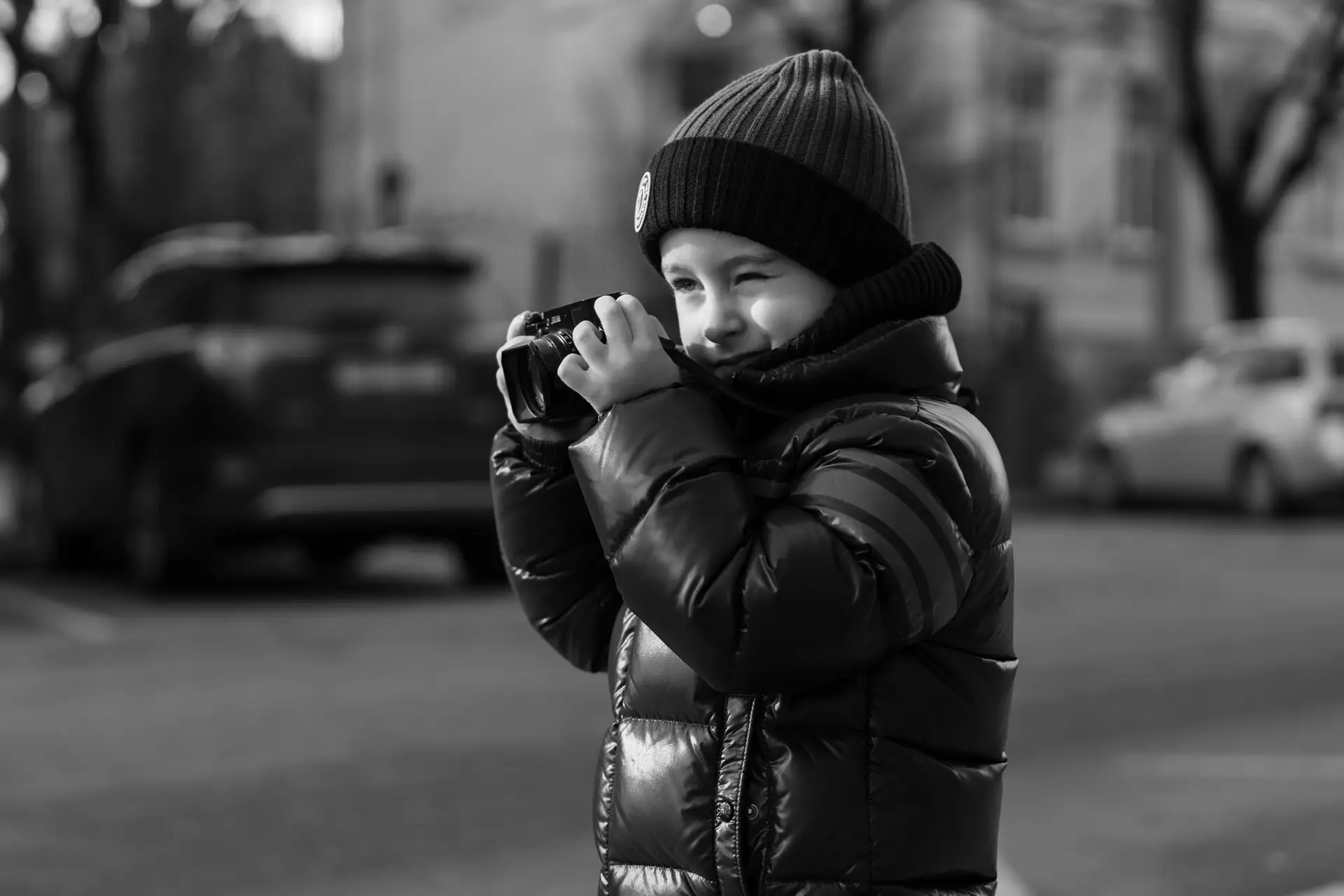 Young boy in a winter jacket and beanie smiling while holding a camera and taking a photo, captured in black and white.