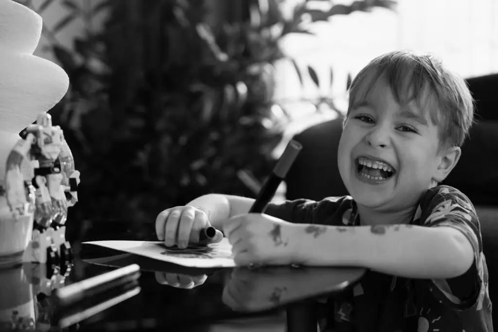 Laughing young boy drawing at a table with markers, captured in a candid black and white photo