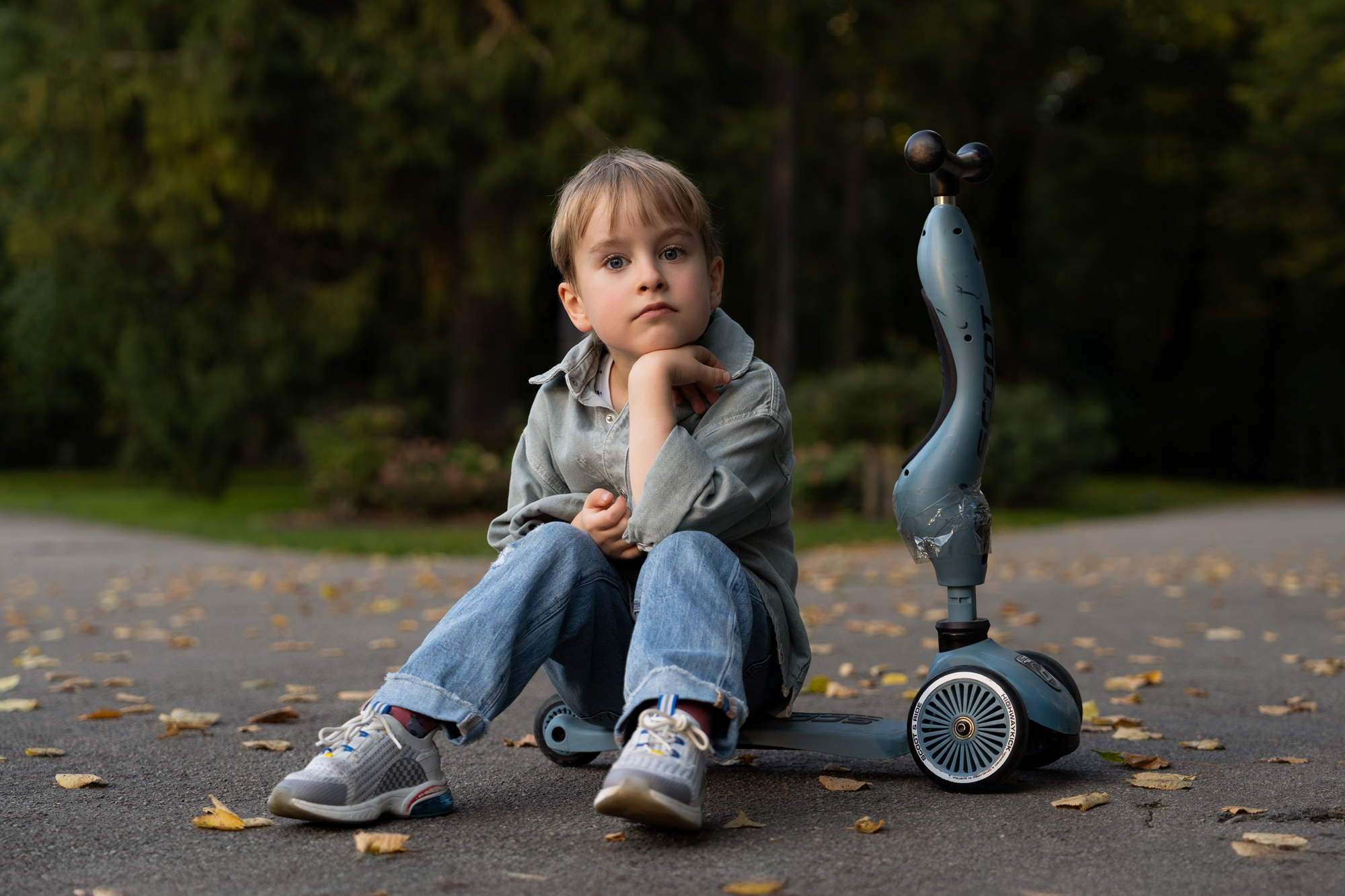 Young boy sitting thoughtfully on the ground next to his scooter in a park with autumn leaves around him.