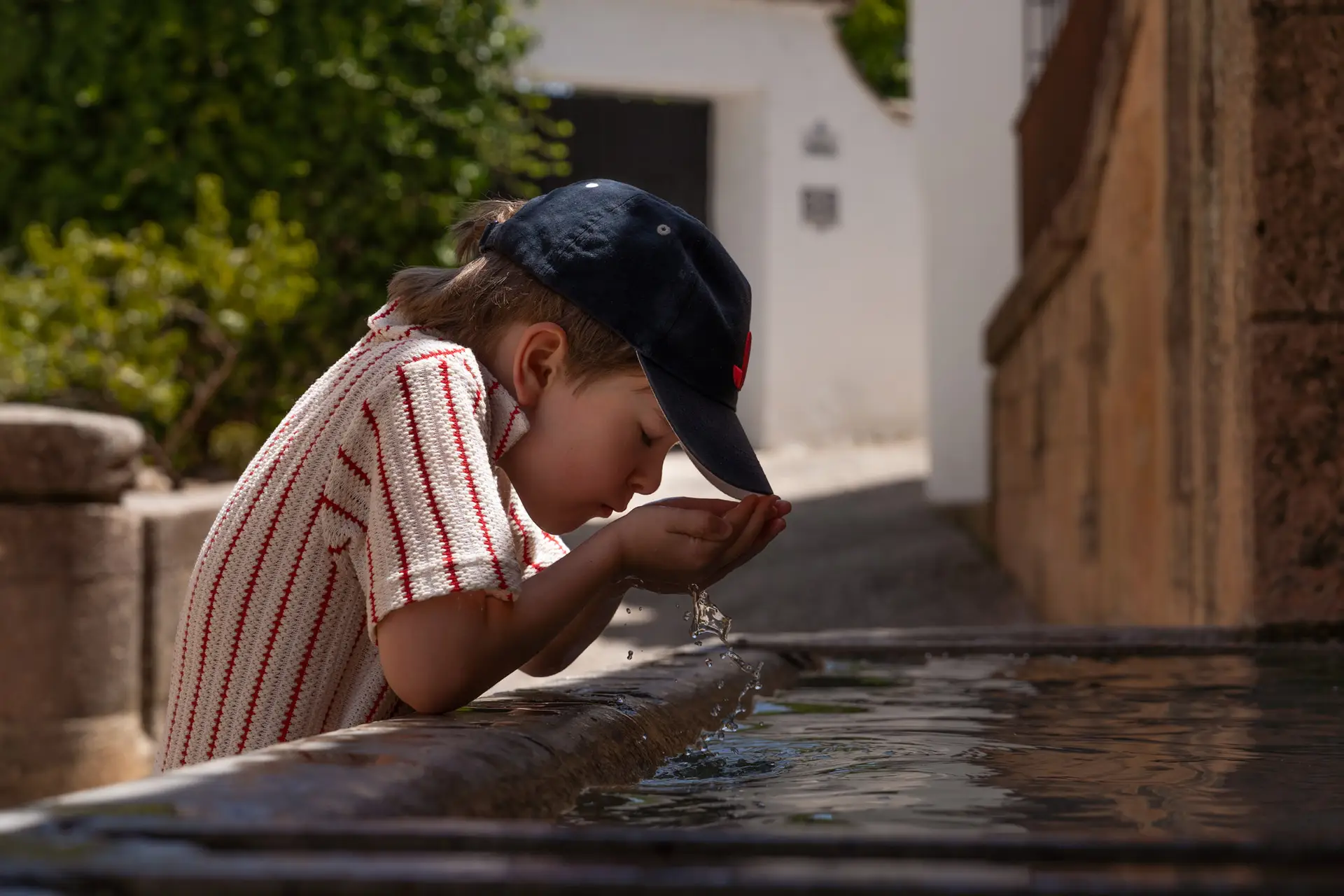 Young boy in a cap drinking water from a stone fountain during a family vacation abroad.