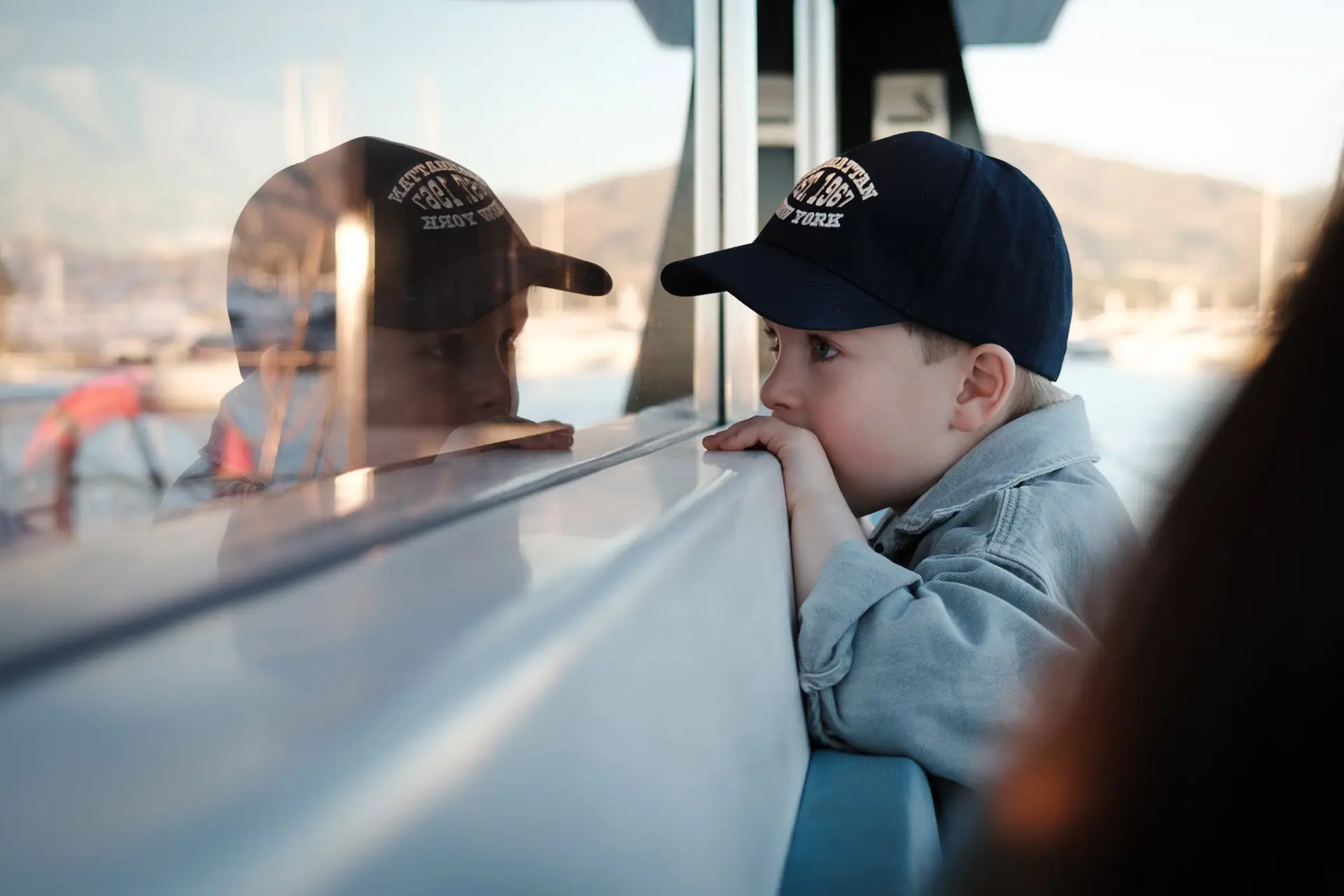 Young boy in a cap leaning on a boat window and looking at his reflection during a family vacation.