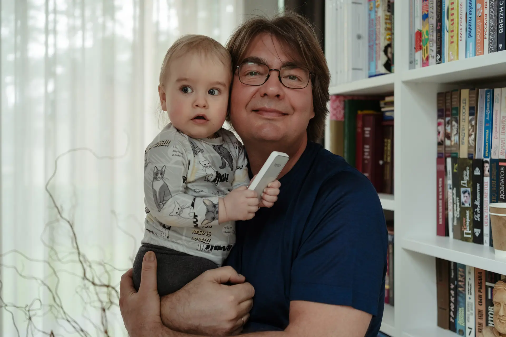 Grandfather holding his grandson indoors, family photography with natural light by the bookshelf.