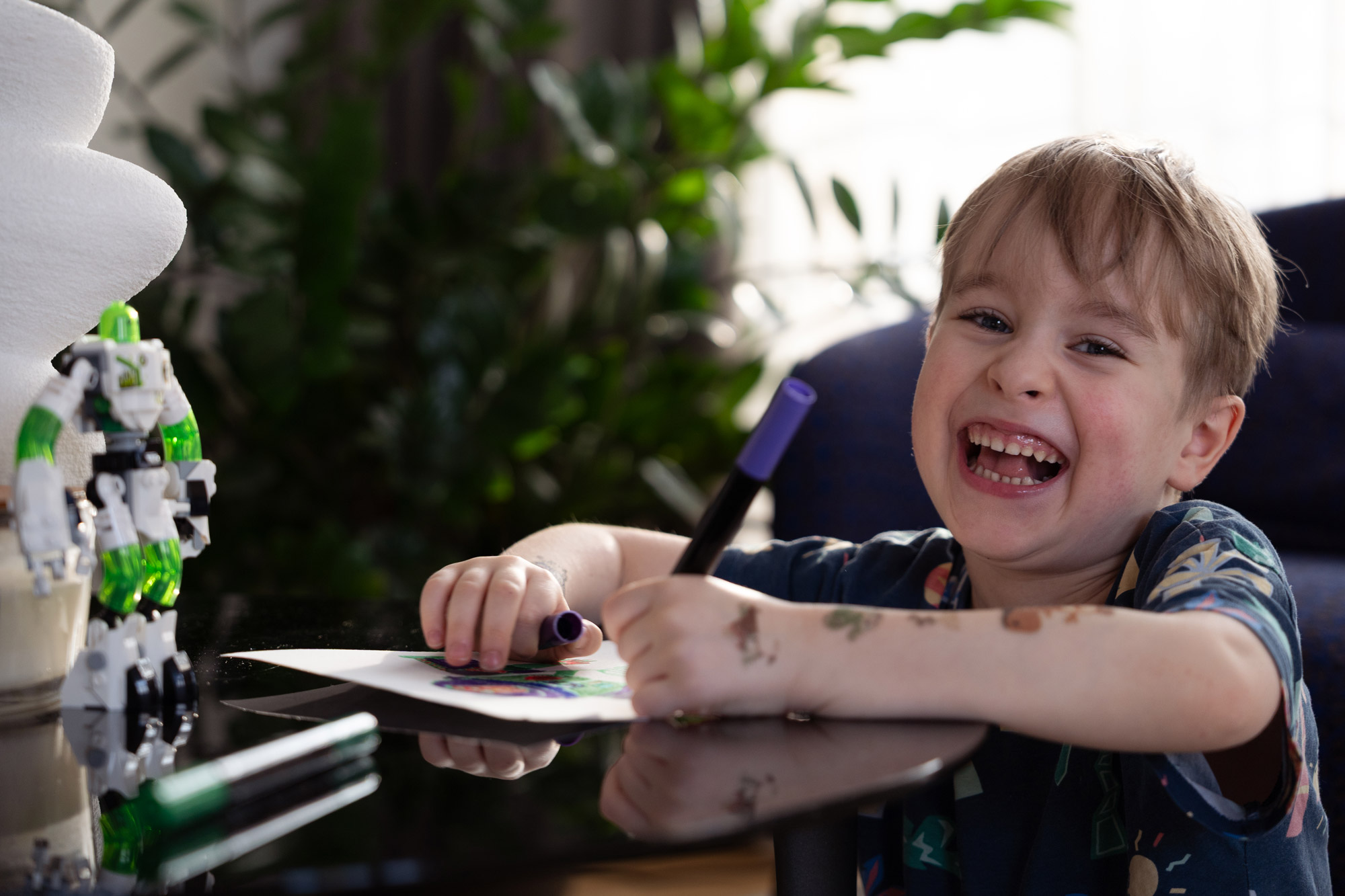 Child laughing while drawing at a table with natural backlighting in indoor family photography.
