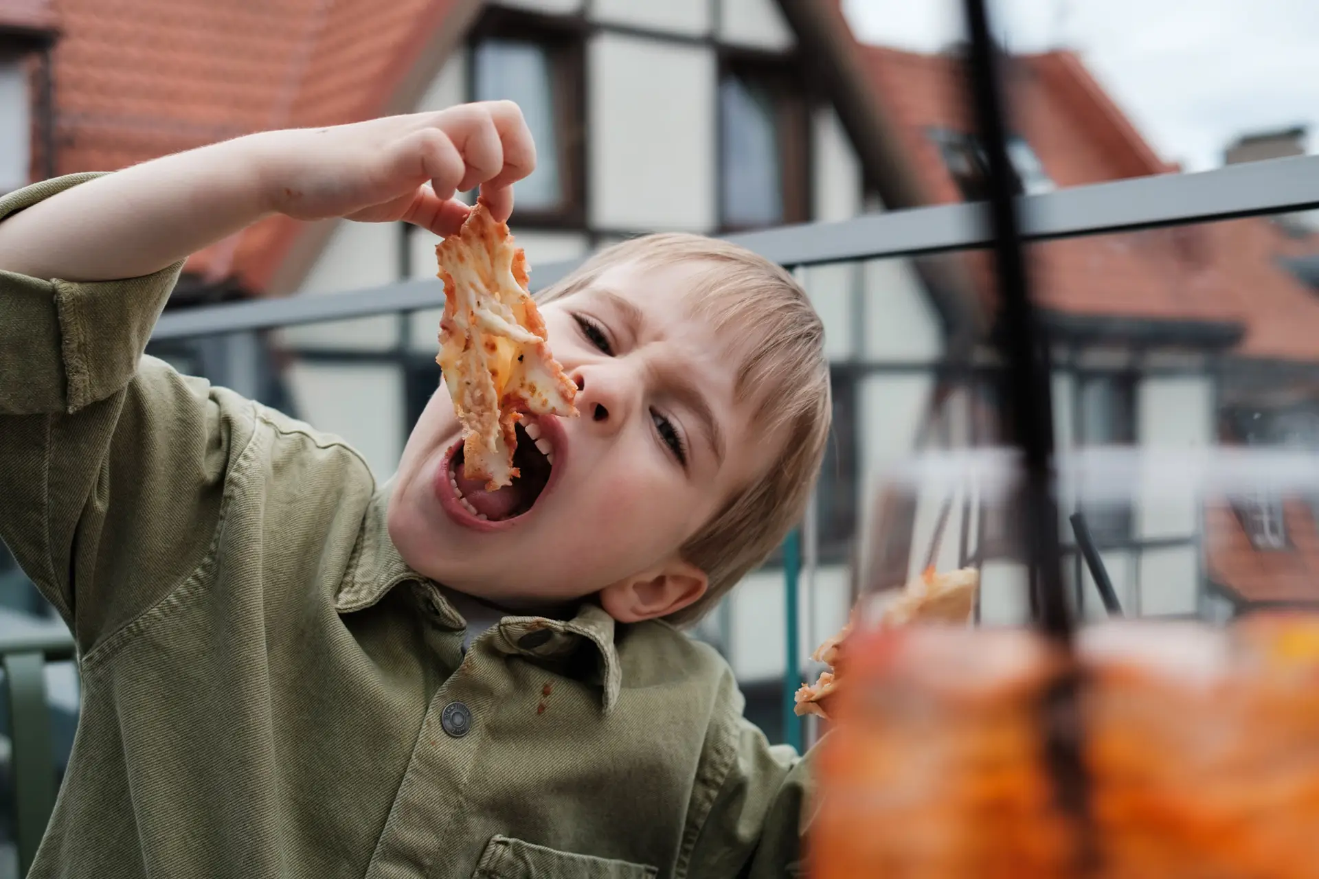 Young boy playfully eating pizza outdoors, holding up a messy slice with sauce on his face and shirt.