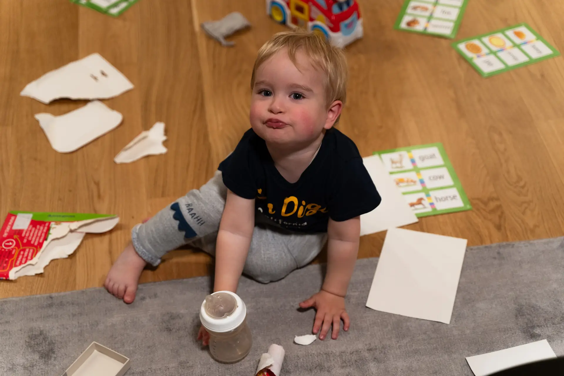oddler sitting on the floor with a milk bottle, surrounded by torn paper and toys, looking up with a playful expression.